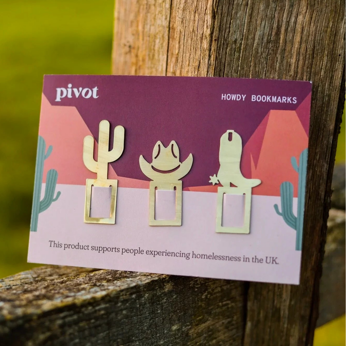 Three small brass bookmarks shaped like a cactus, a cowboy hat, and a cactus are attached to a product card labeled 'Pivot' and 'HOWDY BOOKMARKS', leaning against a weathered wooden post outdoors with blurred green grass in the background.