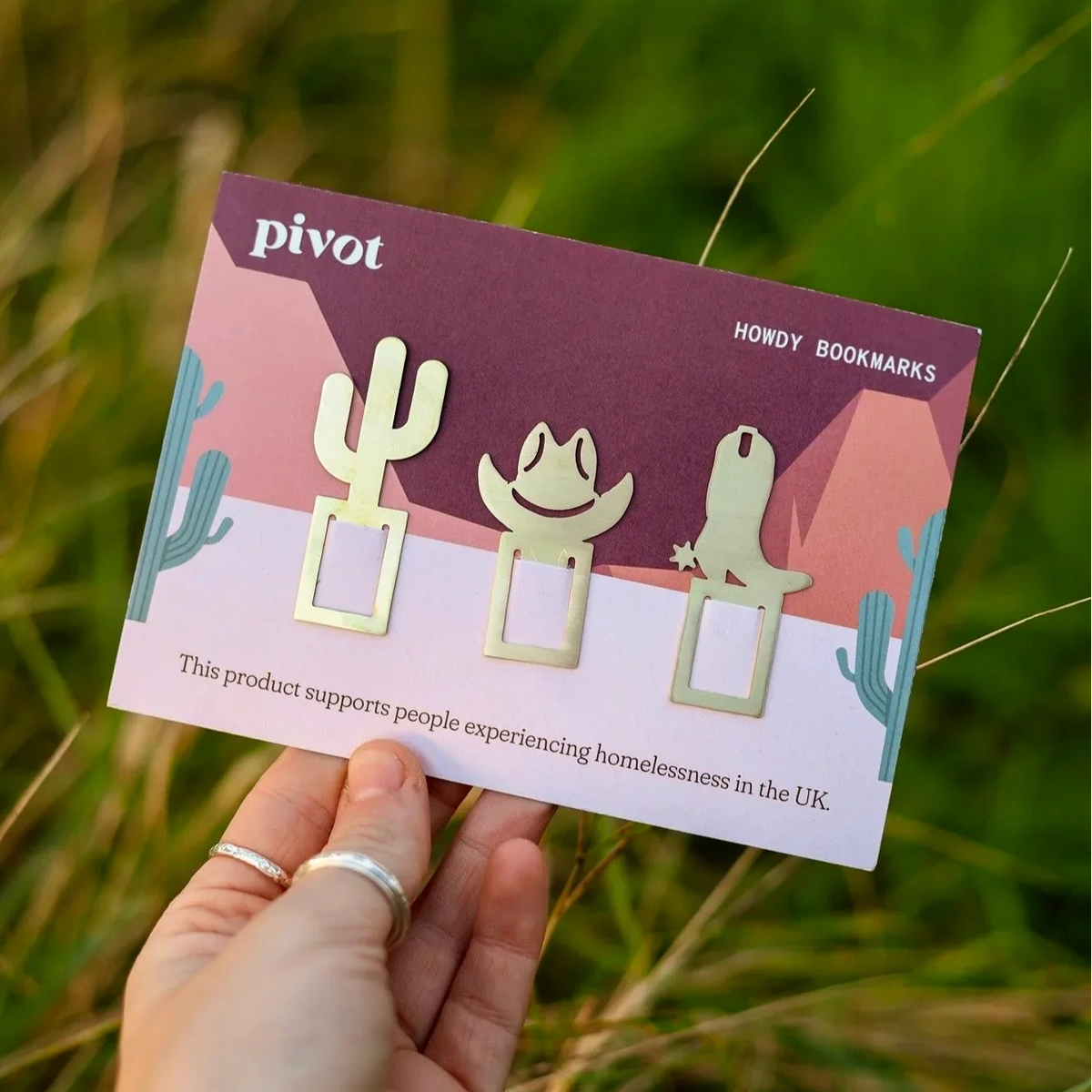 A hand holds a card displaying three brass metal bookmarks shaped like a cactus, a cowboy hat, and a cowboy boot, with a blurred green grassy background.