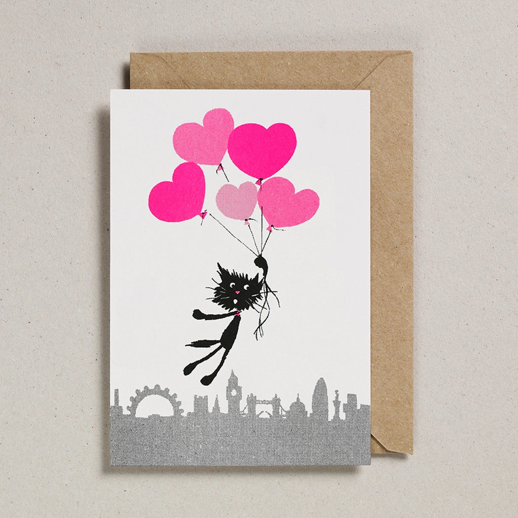 White card with a black and white cat floating above the London skyline holding fluoro pink heart shaped balloons.  With a brown envelope on a white background.