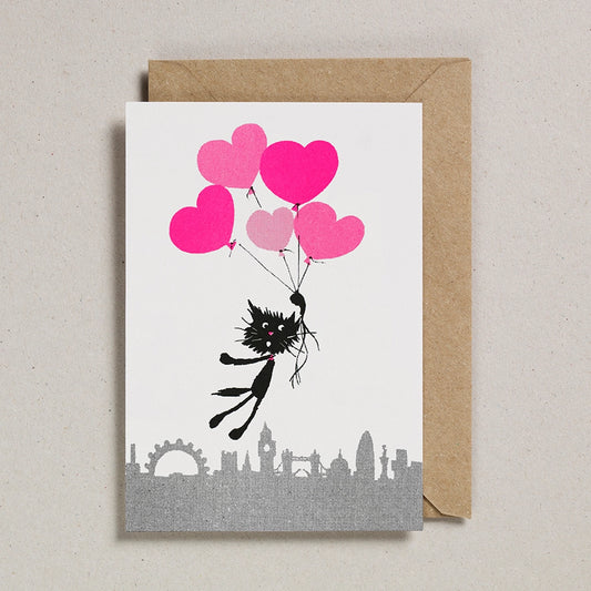White card with a black and white cat floating above the London skyline holding fluoro pink heart shaped balloons.  With a brown envelope on a white background.