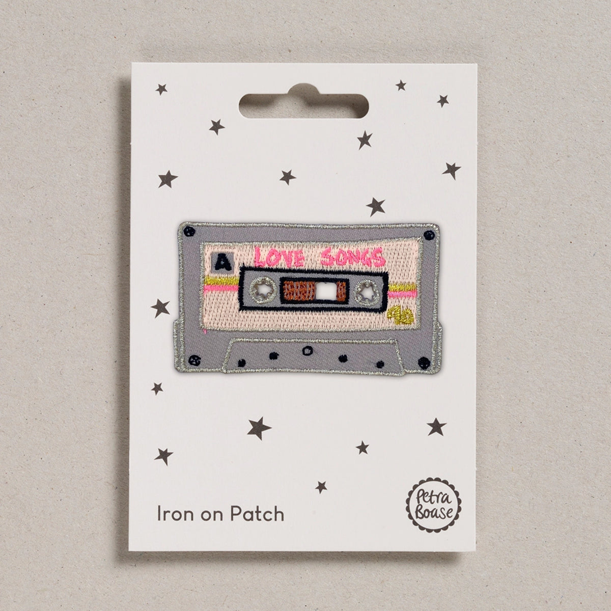 an embroidered patch of a Love Songs mix tape, with pink and gold details. Backed on a white card with black stars.