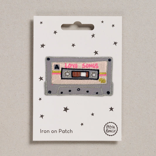 an embroidered patch of a Love Songs mix tape, with pink and gold details. Backed on a white card with black stars.