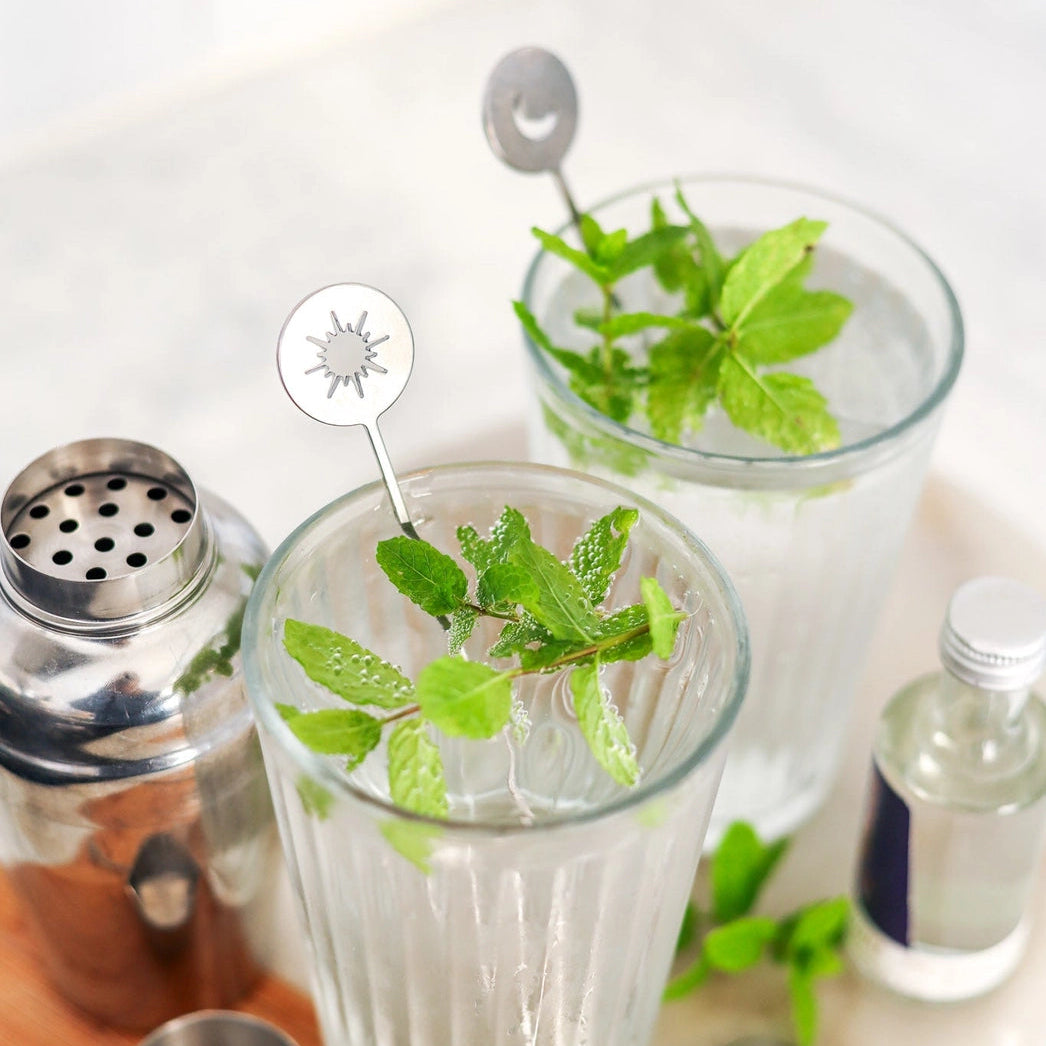 Two clear glass tumblers filled with a clear drink and fresh mint leaves, styled on a wooden board with a metal shaker, strainers, and a small bottle on a light marble surface.