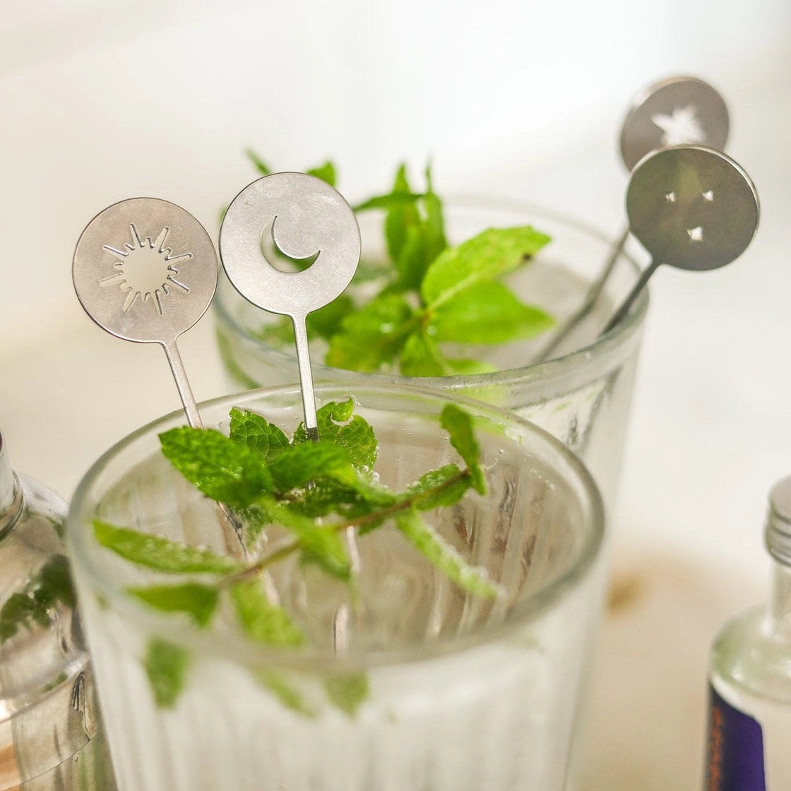 Two clear ribbed drinking glasses filled with a pale beverage and garnished with fresh mint leaves, each holding a decorative metal cocktail stirrer with a moon , star or sun design, set against a softly blurred light background.