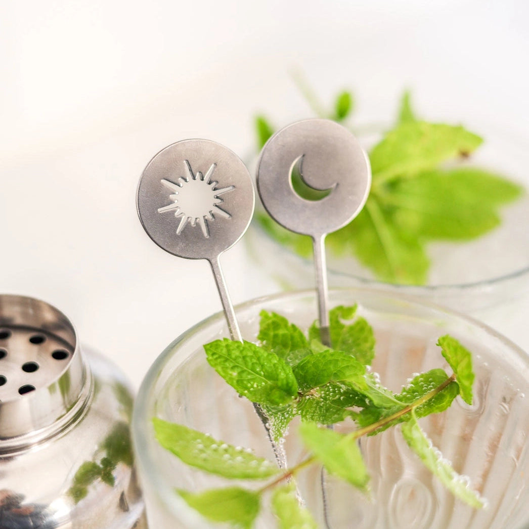 Two silver metal cocktail straws with sun and moon cutouts, displayed in a clear glass filled with fresh mint leaves, alongside a metal shaker.