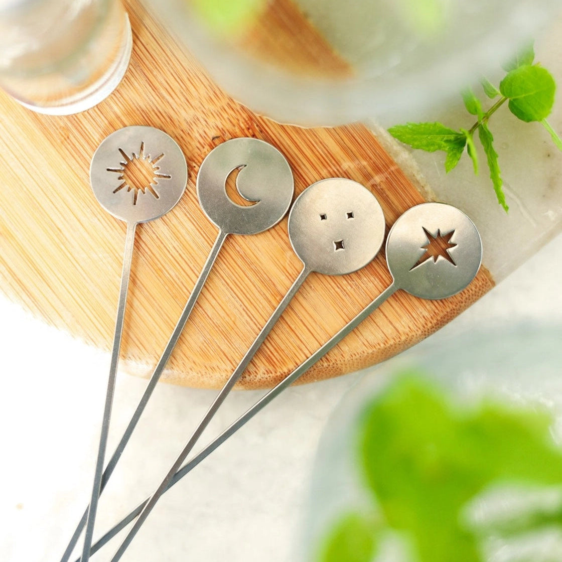 Four metal cocktail straws with circular star, moon, and face cutouts resting on a wooden board, with blurred mint leaves and glassware in the background.