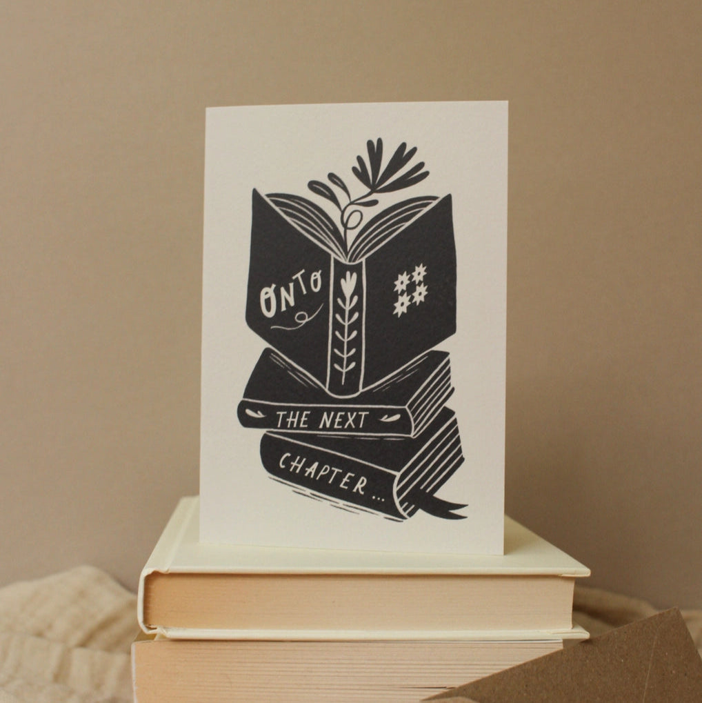 Greeting card with 'Onto the Next Chapter'  book stack design on a beige background with a brown envelope, sat on top of a stack of beige books.