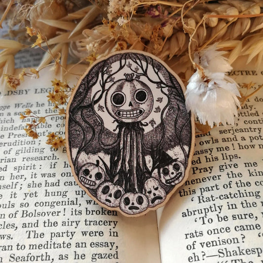 Wooden pin brooch with a decorative black and white, jack'o'lantern design, featuring pumpkins, vines and moonlight. Sat on an open book.