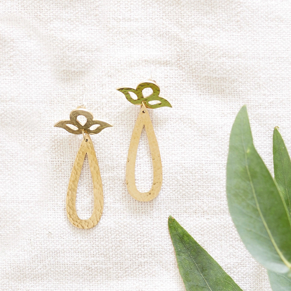 Hammered brass drop studs in a peacock design on a hessian background with green leaves.