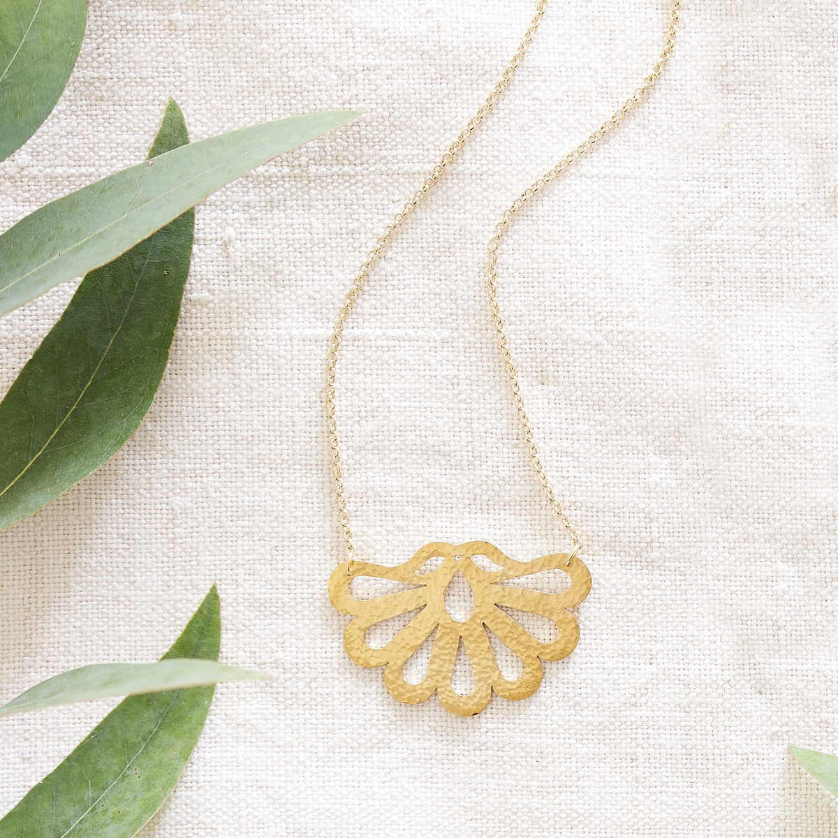 Hammered brass peacock feather necklace with chain, on a hessian background with green leaves.