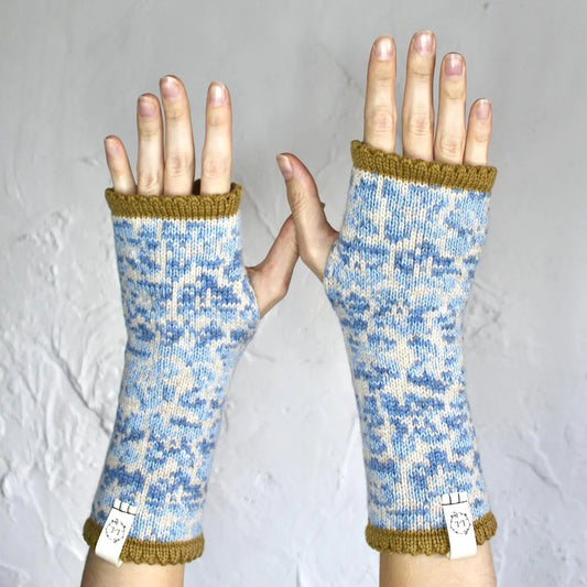 A pair of fingerless knit gloves with a blue and white floral pattern and mustard yellow trim, shown on a person's hands against a textured gray background.