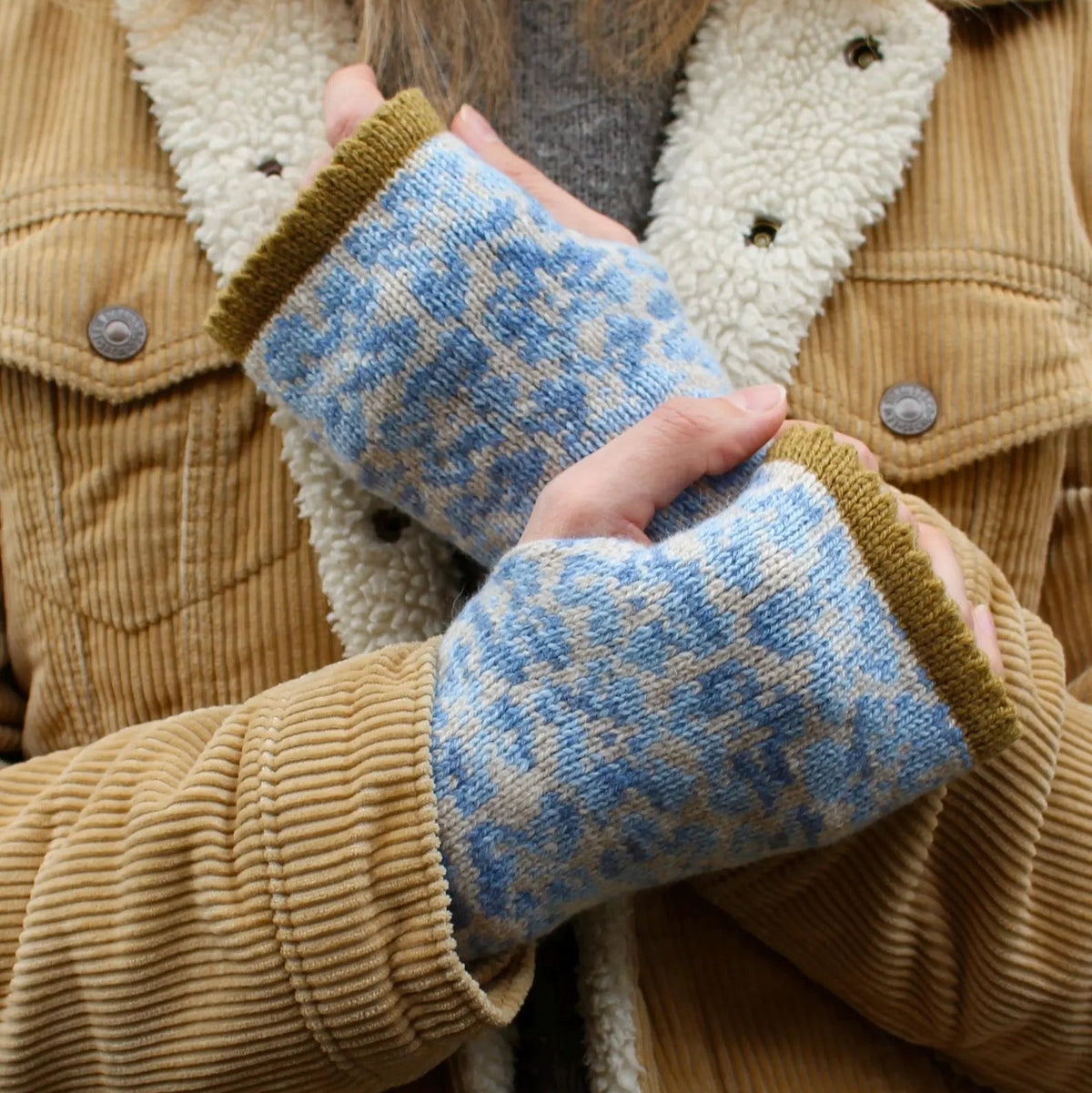 Close-up of a person wearing fingerless knit gloves with a blue and beige floral pattern and mustard yellow ribbed cuffs, shown over a tan corduroy jacket with cream sherpa lining.