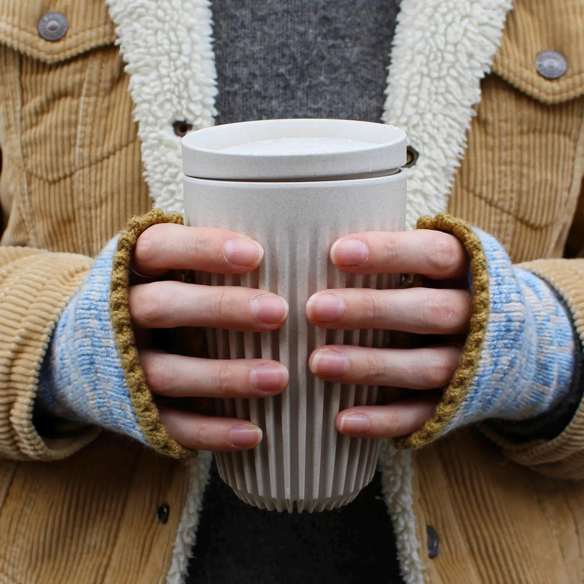 A close-up of a person holding a white ribbed paper coffee cup with a lid, wearing a tan corduroy jacket with a cream sherpa lining, a gray sweater, and blue knit fingerless gloves.
