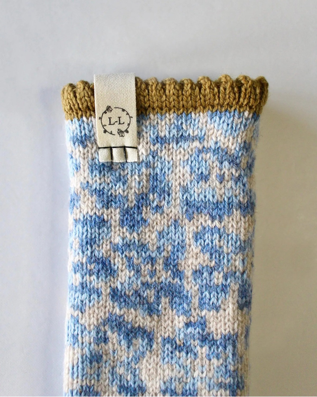 A close-up of a knitted glove cuff with a blue and off-white floral pattern, finished with a mustard yellow ribbed top edge and a white cotton brand label attached.