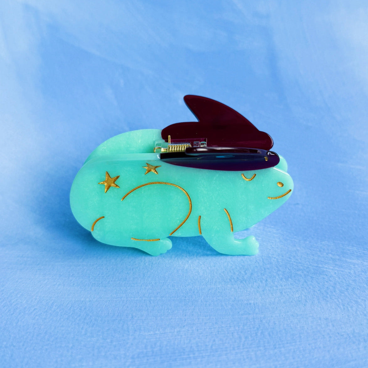 Pale green toad design hair claw with red witches hat on pale blue background.
