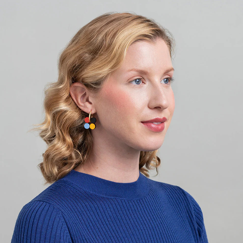 A person with wavy blonde hair wearing a blue sweater and colorful flower-shaped earrings looks to the side against a plain background.