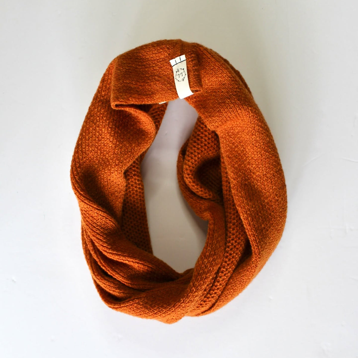 An orange knit infinity scarf with a small white brand tag, displayed on a clean white background.