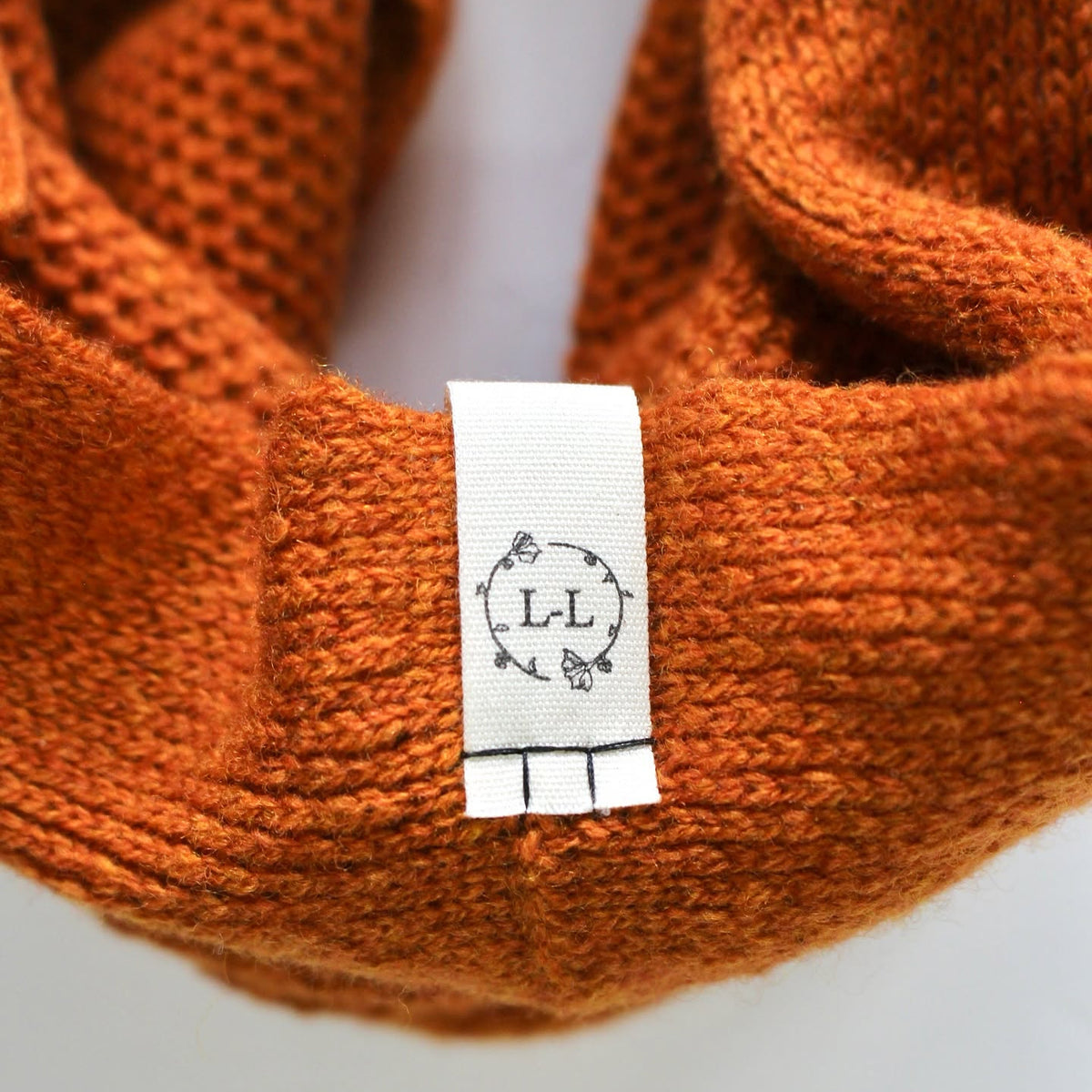 Close-up of a textured orange knit infinity scarf with a white fabric size tag showing 'L-L' in a circular design.