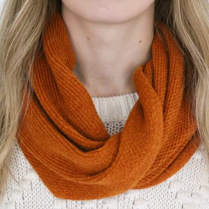 A close-up of a woman wearing a thick, ribbed infinity scarf in a warm burnt orange color, paired with a white knit sweater.