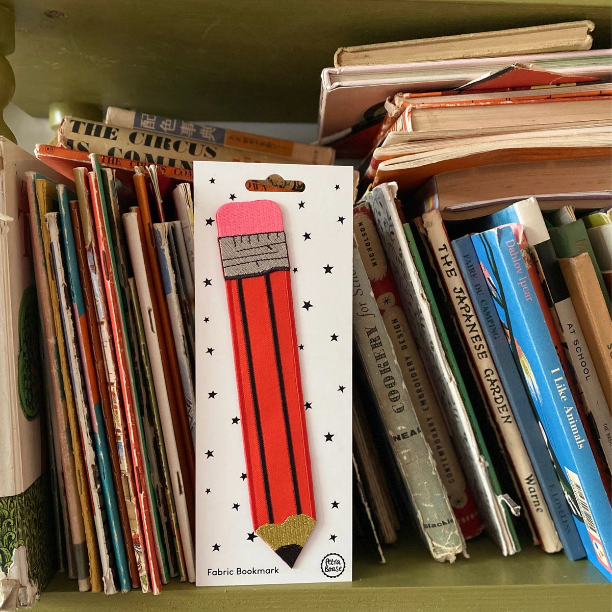 Fabric bookmark with red and gray design in a box, surrounded by books on a shelf.
