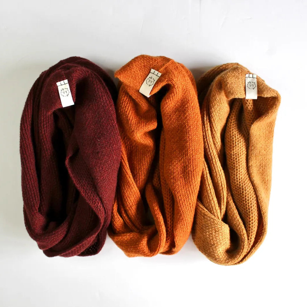 Three folded knit infinity scarves in burgundy, burnt orange, and camel brown, arranged side-by-side on a white background.