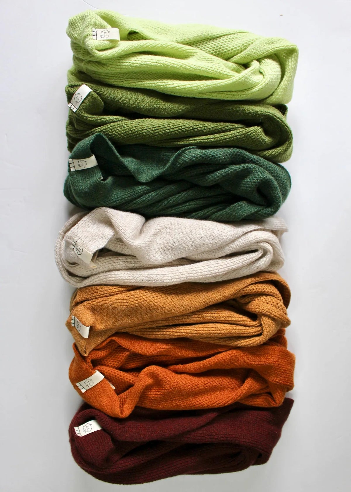 A vertical stack of eight folded textured knit scarves in various earthy and vibrant colors, including lime green, olive green, forest green, cream, mustard, orange, and burgundy, on a plain white background.