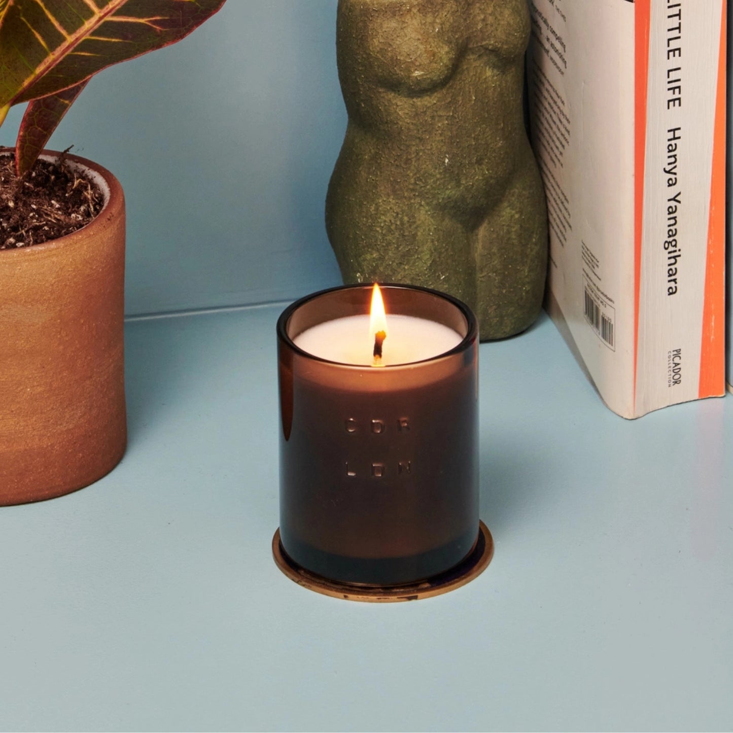 Candle in a brown glass jar on a light blue surface with a plant and book in the background