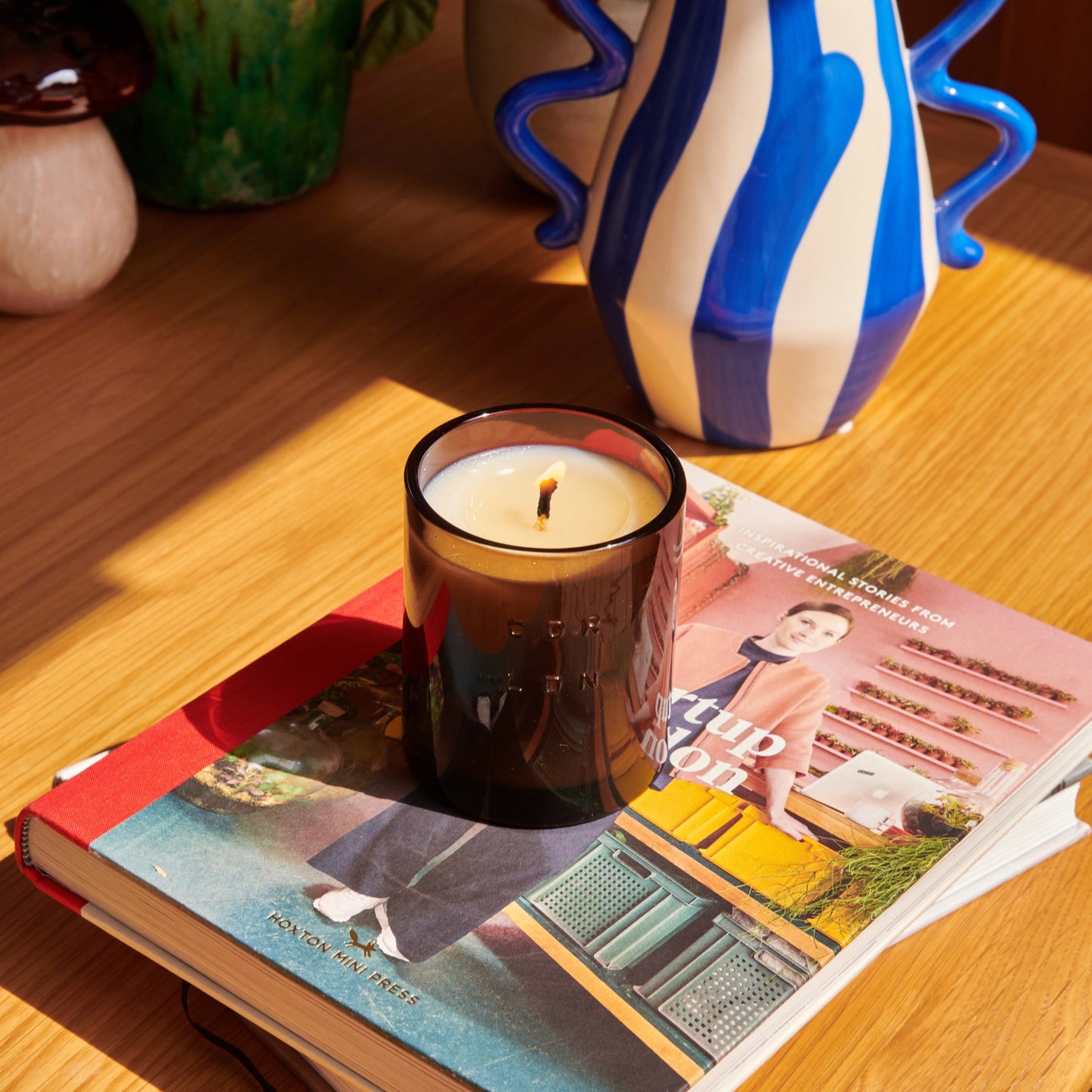 Candle in an amber jar on an open book with decorative vases in the background.