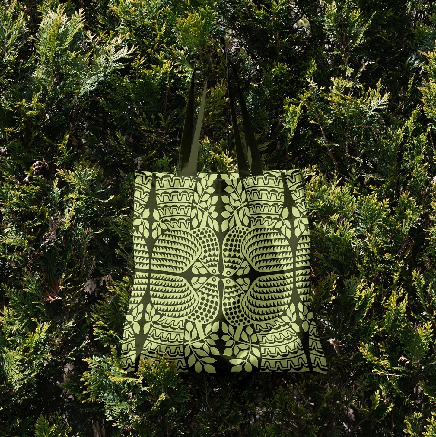 Green Peacock design shopper tote bag hangng from a conifer bush. Storigraphic & Hornsea pottery collaboration.