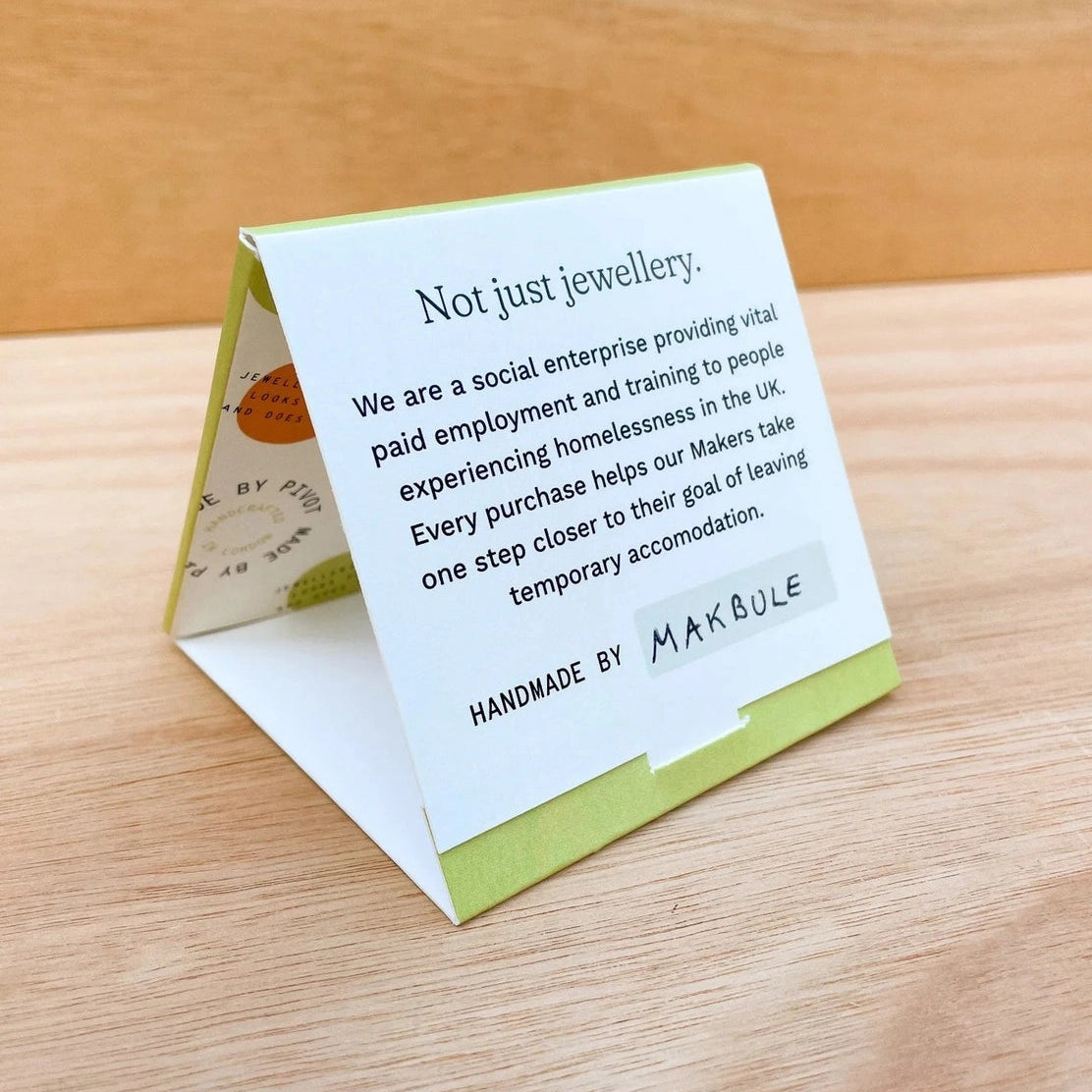 A small folded paper sign or card on a wooden tabletop, printed with text about Pivot the social enterprise and the makers' name Makbule. The card has a white background with green border accents and is photographed in a real indoor setting.
