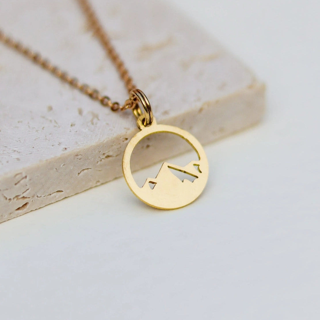 A gold necklace with a circular pendant featuring a minimalist mountain silhouette, displayed on a light beige stone slab against a white background.
