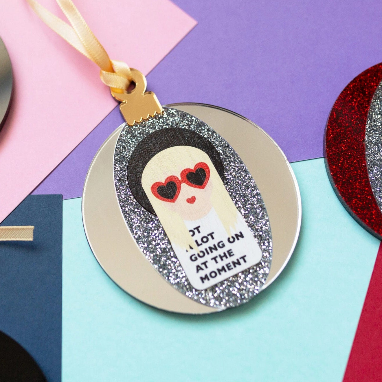Silver & mirror Taylor Swift acrylic bauble, on a muclticoloured background.