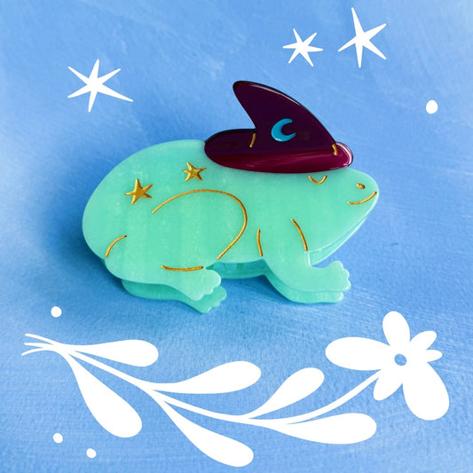 Green frog-shaped 9cm hair claw with a purple witch hat on a blue background with white stars and floral elements.