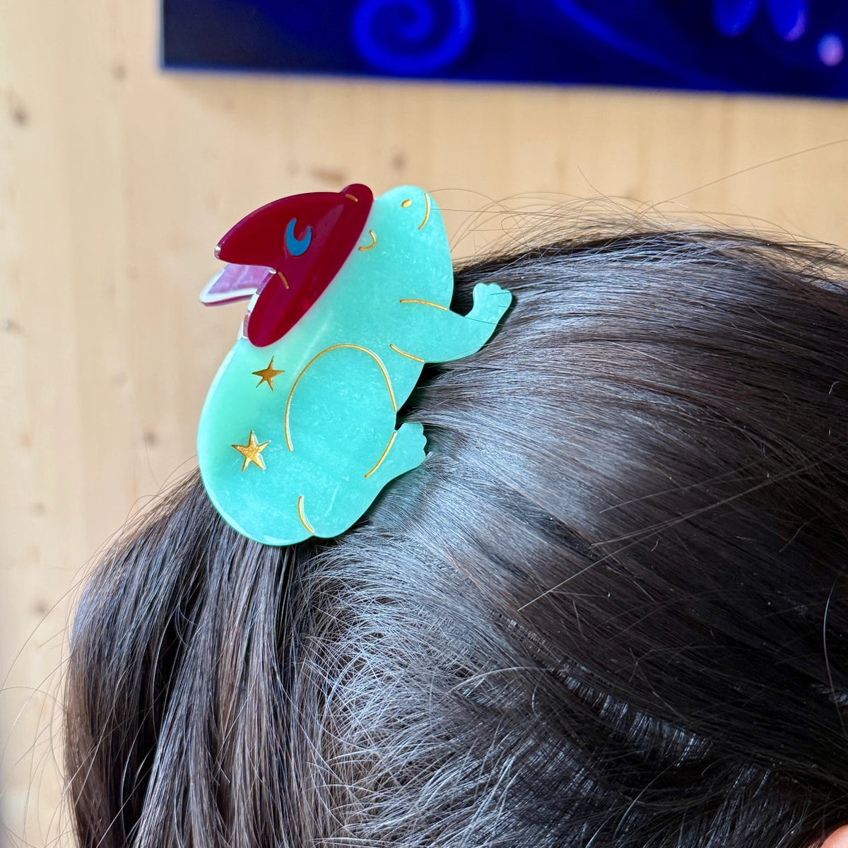 Turquoise toad hair clip with red and yellow accents on a person's head against a neutral background.