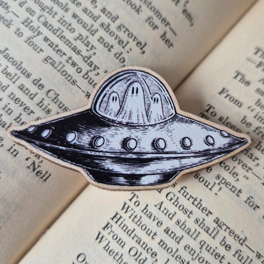 A woonden pin badge with a black and white design, featuring a flying UFO with ghosts inside.